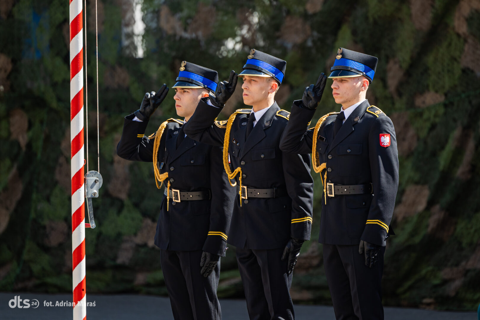 Three uniformed soldiers salute beside a striped pole during a ceremony, Poland patch visible on one sleeve.