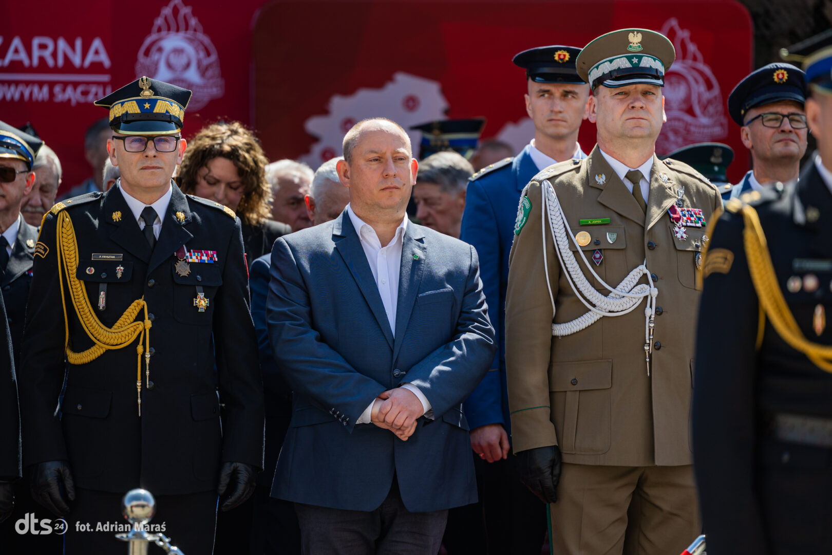 Group of uniformed military officers standing at a formal ceremony, with a man in a blue suit centered among them.