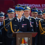 Uniformed fire service officer speaks at a podium during a formal ceremony, with colleagues in dress uniforms behind him.