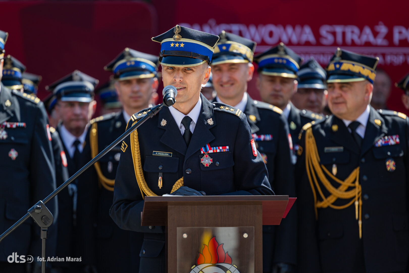 Uniformed fire service officer speaks at a podium during a formal ceremony, with colleagues in dress uniforms behind him.