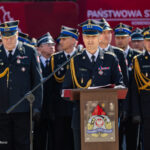 Central officer in ceremonial uniform speaks at a podium during a fire service ceremony, surrounded by colleagues in dress uniform with gold cords and insignia.