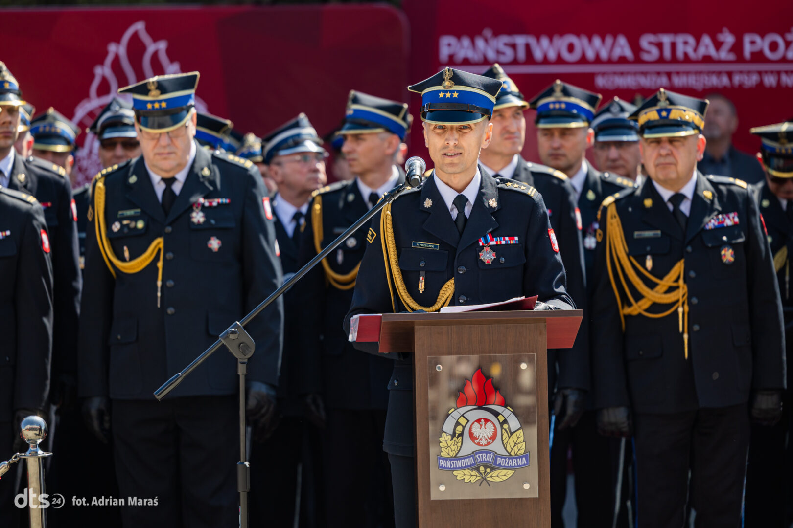 Central officer in ceremonial uniform speaks at a podium during a fire service ceremony, surrounded by colleagues in dress uniform with gold cords and insignia.