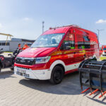 Red fire brigade van with STRAŻ markings in a paved industrial yard, with an all-terrain motorcycle nearby and crew nearby.