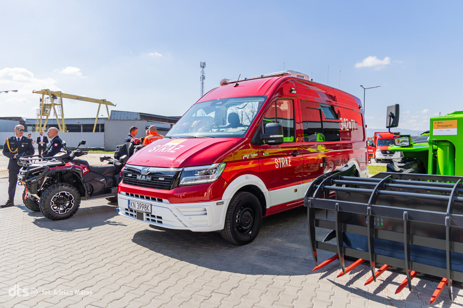Red fire brigade van with STRAŻ markings in a paved industrial yard, with an all-terrain motorcycle nearby and crew nearby.