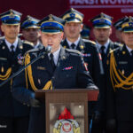 Officer in formal military uniform speaks at a podium during a ceremony, with uniformed colleagues behind him.