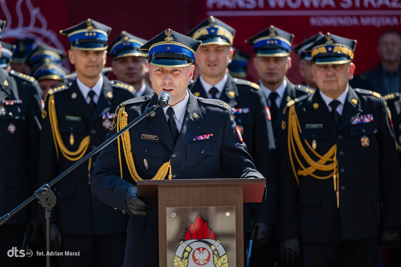 Officer in formal military uniform speaks at a podium during a ceremony, with uniformed colleagues behind him.