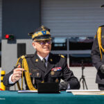 Two military officers in dress uniforms sit and stand at a table with papers outside a building; the seated man gestures with a pen as the other smiles nearby.