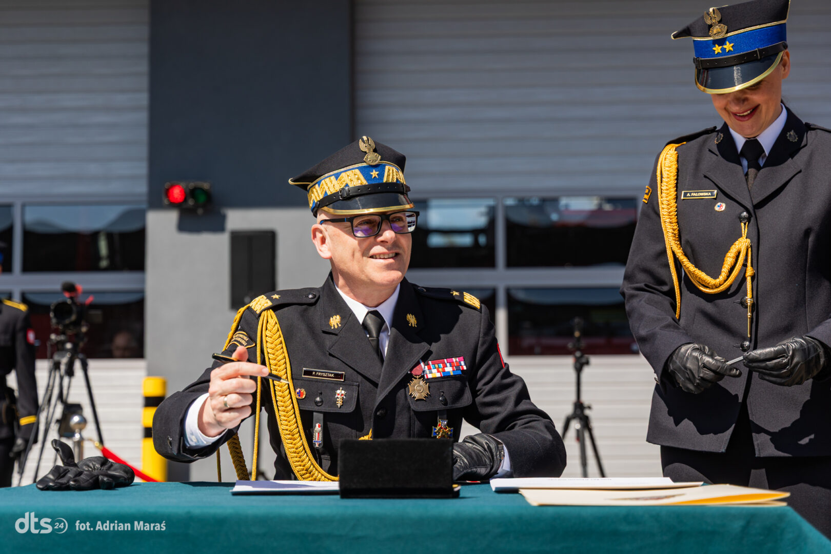 Two military officers in dress uniforms sit and stand at a table with papers outside a building; the seated man gestures with a pen as the other smiles nearby.