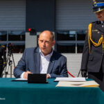Man in a dark suit signing a document at a teal table during a formal ceremony, uniformed officers stand in the background outdoors