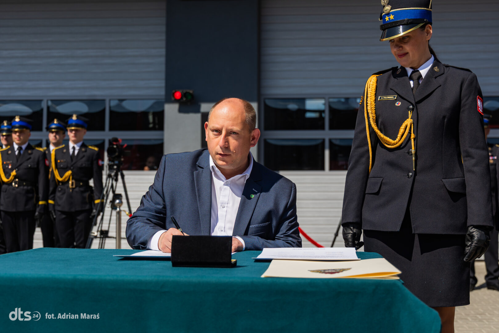 Man in a dark suit signing a document at a teal table during a formal ceremony, uniformed officers stand in the background outdoors
