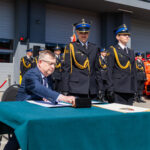 Man in a suit signs documents at a table during a formal ceremony, with uniformed officers standing in formation behind him.