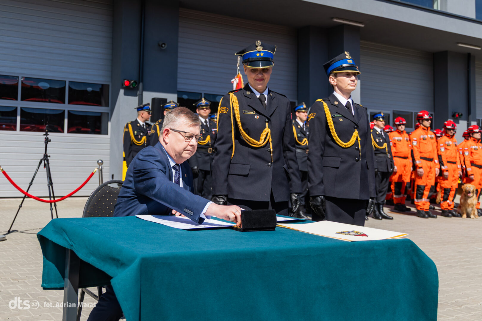 Man in a suit signs documents at a table during a formal ceremony, with uniformed officers standing in formation behind him.
