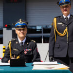 Two uniformed officers at a signing table during a formal military ceremony; one is seated signing papers, the other stands beside.