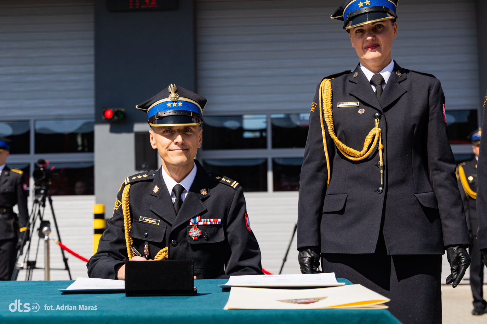 Two uniformed officers at a signing table during a formal military ceremony; one is seated signing papers, the other stands beside.