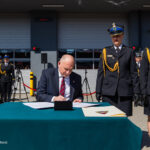 A man in a suit signs a document at a teal table while uniformed officers stand at attention in the background.