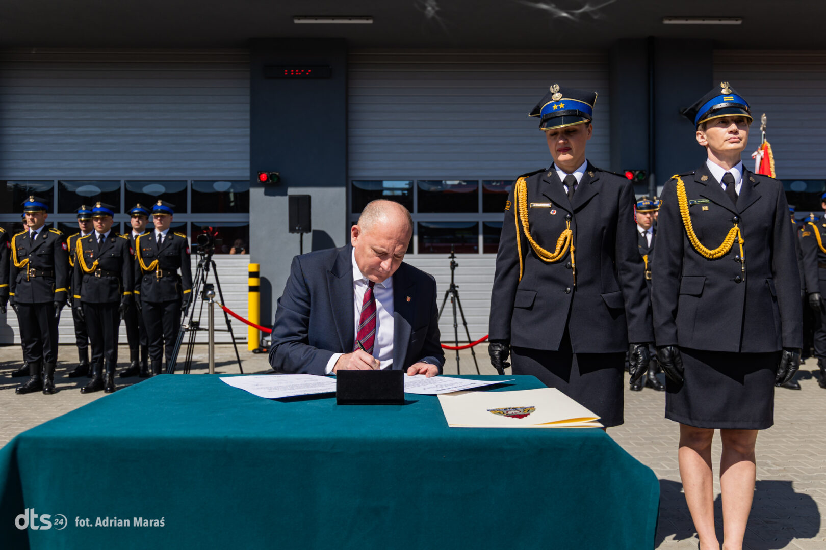 A man in a suit signs a document at a teal table while uniformed officers stand at attention in the background.