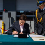 Woman in a dark suit signs a document at a teal table during a formal outdoor ceremony, with a uniformed officer beside her and cameras in the background.