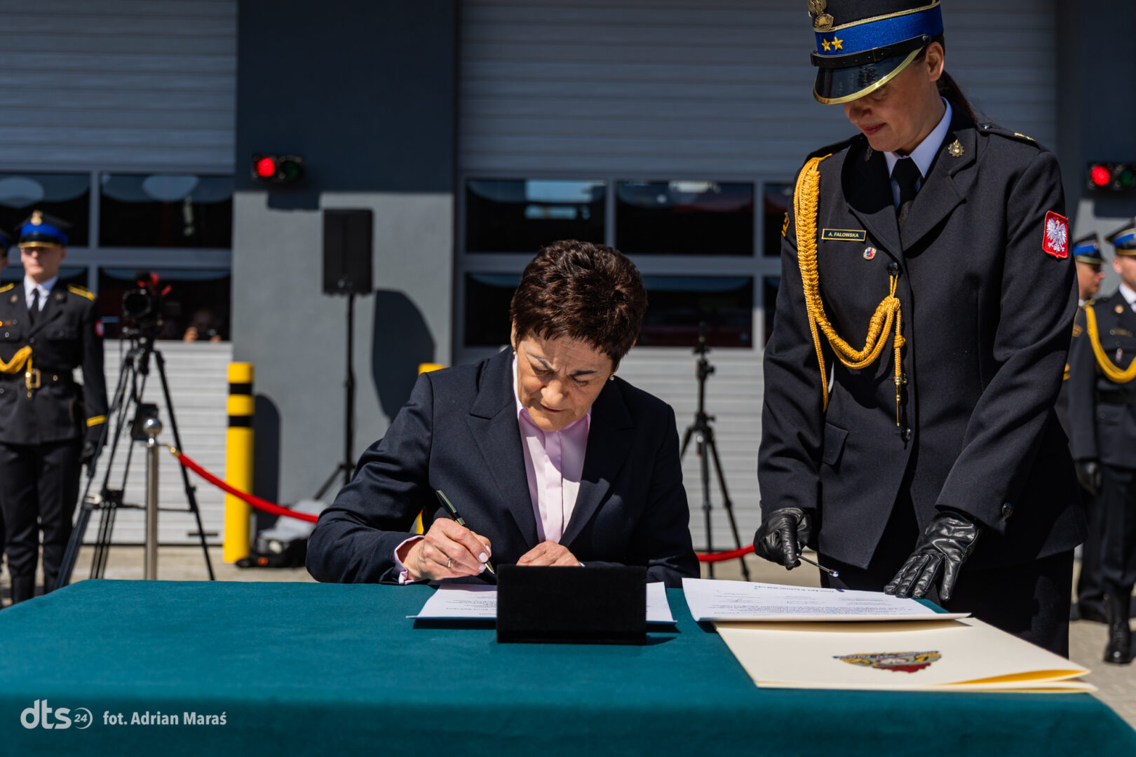 Woman in a dark suit signs a document at a teal table during a formal outdoor ceremony, with a uniformed officer beside her and cameras in the background.