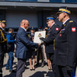 Man in a suit hands a plaque to a Polish military officer during an outdoor award ceremony, with photographers and other uniformed personnel watching.