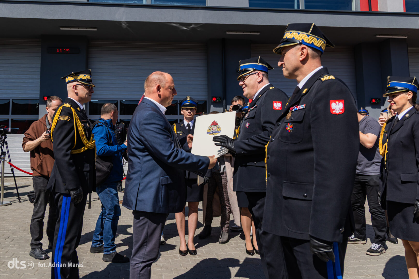 Man in a suit hands a plaque to a Polish military officer during an outdoor award ceremony, with photographers and other uniformed personnel watching.