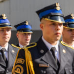 Group of cadets in formal dress uniforms with blue peaked caps and gold trim, standing in formation outdoors during a ceremony