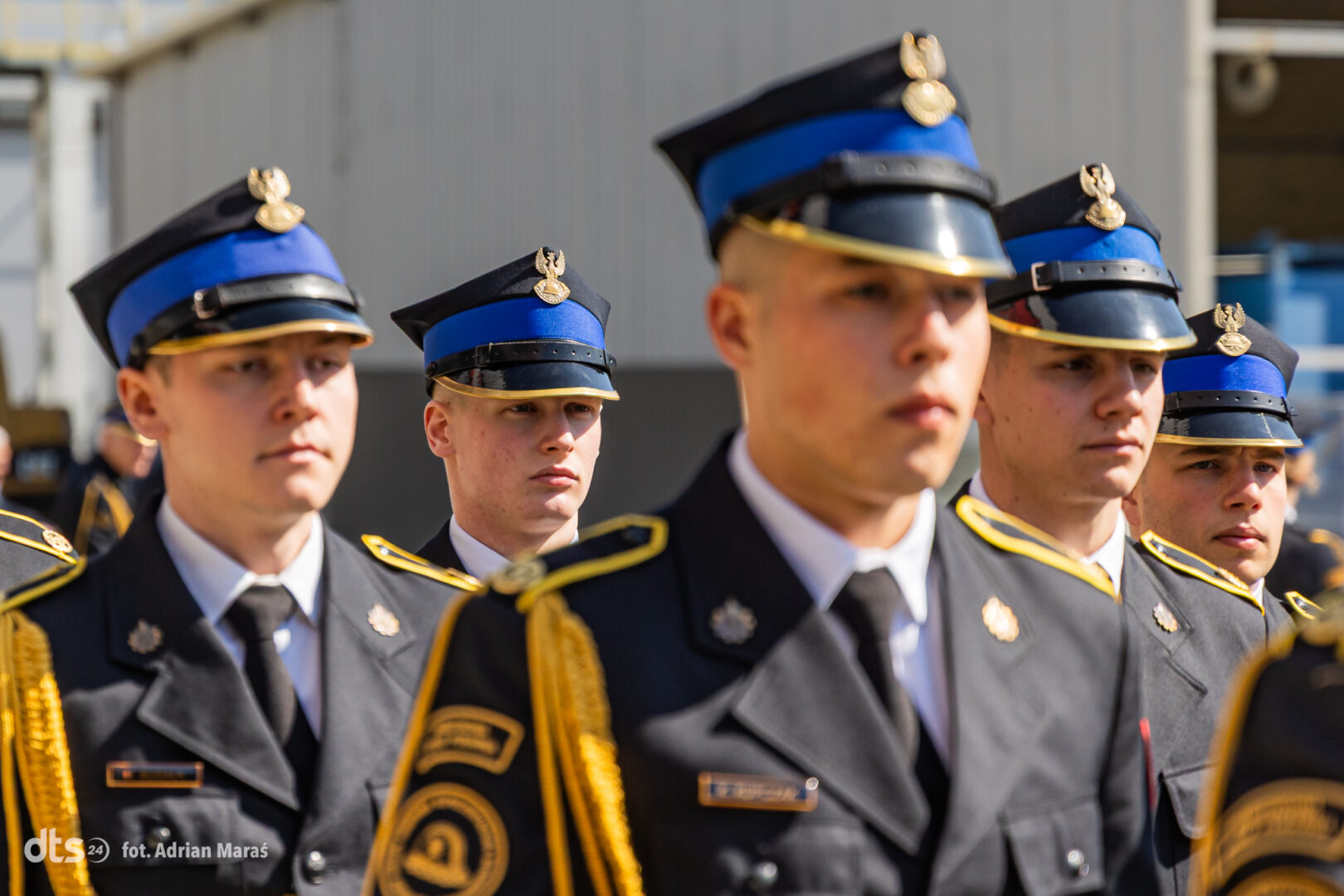 Group of cadets in formal dress uniforms with blue peaked caps and gold trim, standing in formation outdoors during a ceremony