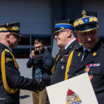 Two Polish military officers in dress uniforms shake hands during a ceremony, medals and gold cords visible, photographer in the background.