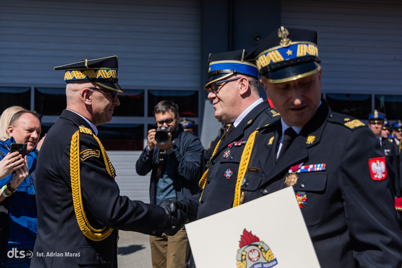 Two Polish military officers in dress uniforms shake hands during a ceremony, medals and gold cords visible, photographer in the background.