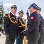 Group of uniformed officers in dress uniforms exchanging a salute outdoors, with a red emergency vehicle in the background.