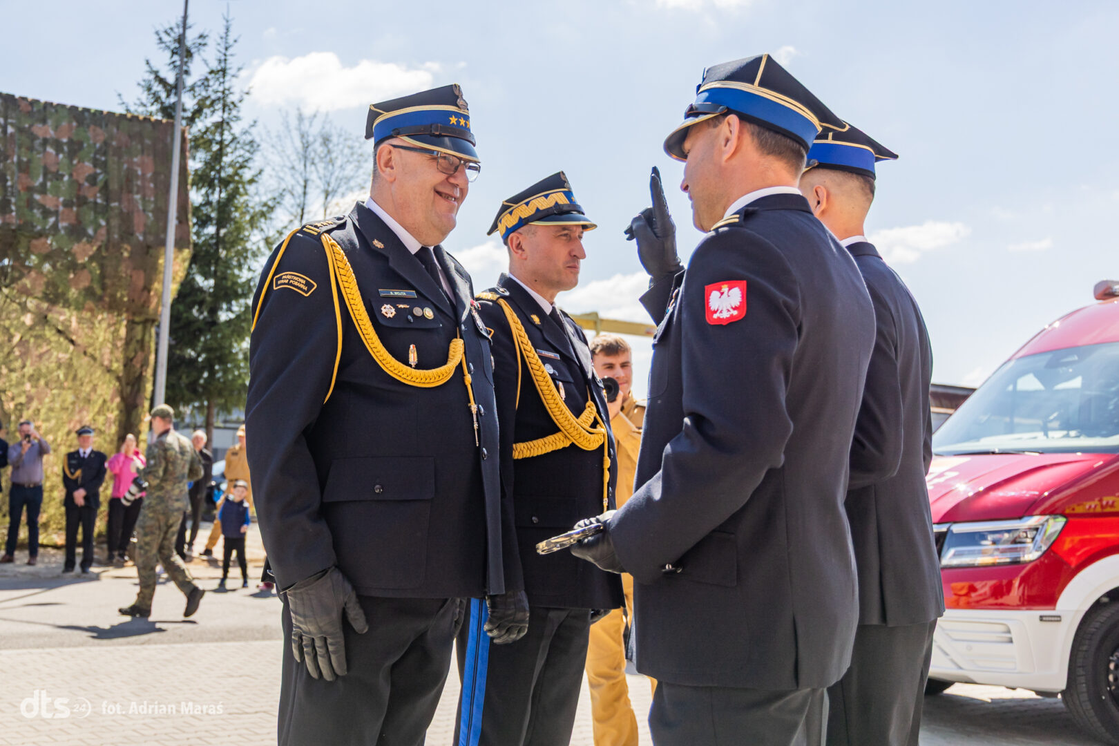 Group of uniformed officers in dress uniforms exchanging a salute outdoors, with a red emergency vehicle in the background.