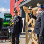 Three Polish military officers in dress uniforms stand outdoors beside a wooden bell frame and industrial equipment.