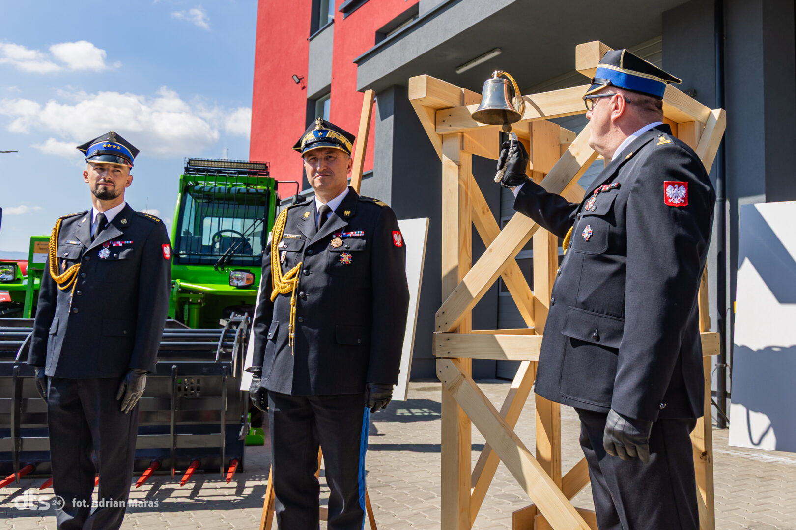 Three Polish military officers in dress uniforms stand outdoors beside a wooden bell frame and industrial equipment.