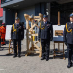 Four uniformed officers stand in a line during a ceremony outside a modern building, with a wooden frame and plaque behind them.