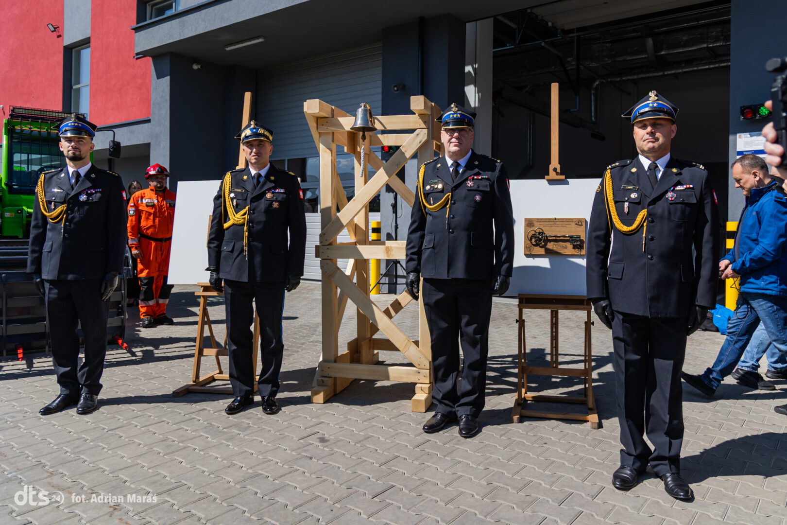 Four uniformed officers stand in a line during a ceremony outside a modern building, with a wooden frame and plaque behind them.