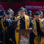 Ceremonial gathering of firefighters with a priest at a podium, wearing ceremonial sashes and uniforms under a red banner.