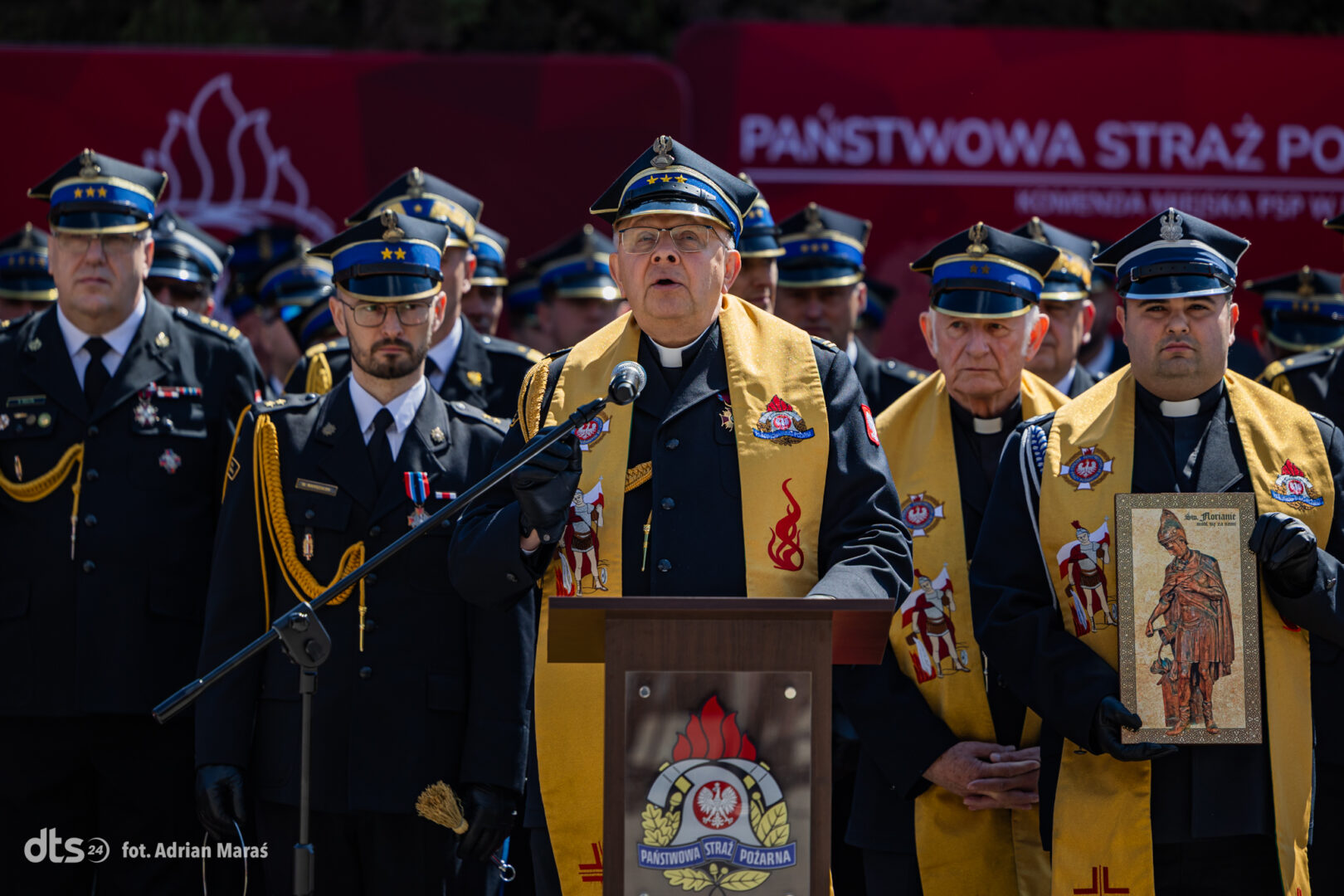 Ceremonial gathering of firefighters with a priest at a podium, wearing ceremonial sashes and uniforms under a red banner.