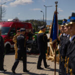 Uniformed personnel line up for a ceremony as a flag bearer presents a blue banner; a red emergency vehicle is parked in the background.