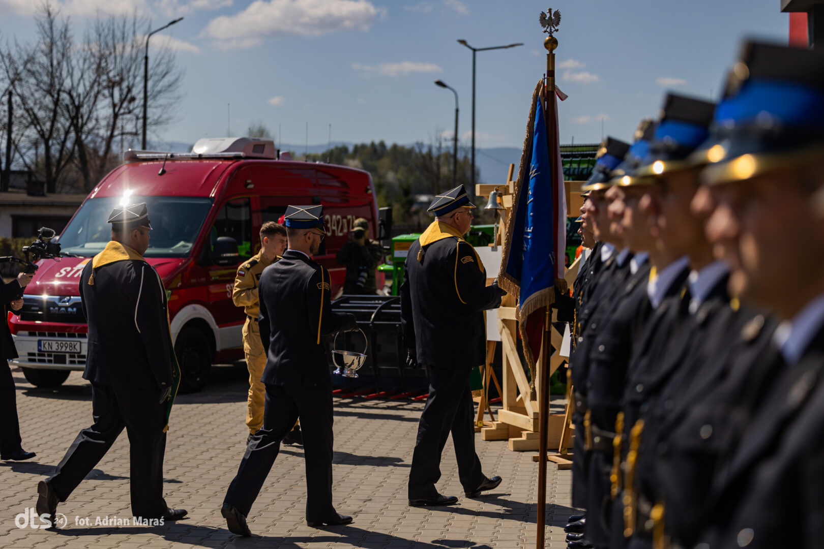 Uniformed personnel line up for a ceremony as a flag bearer presents a blue banner; a red emergency vehicle is parked in the background.