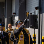 Senior military officer in ceremonial uniform raises a brush, sprinkling particles during a formal outdoor event, with fellow officers and a photographer in the background.