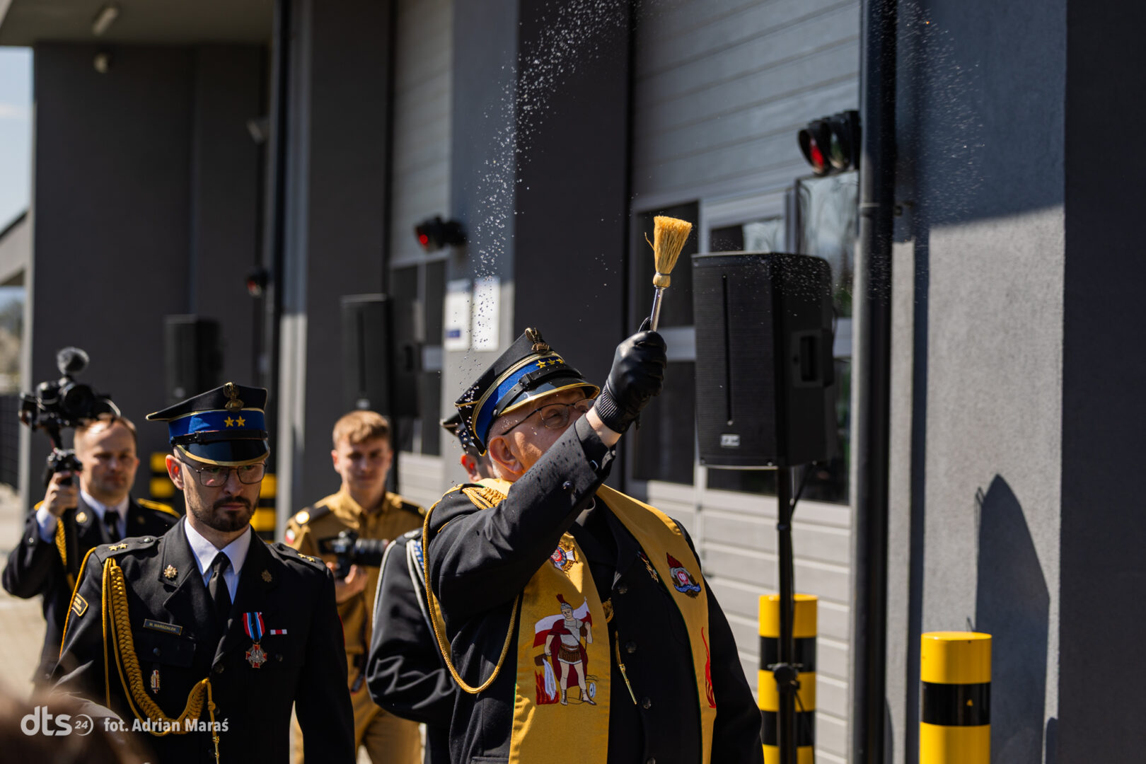 Senior military officer in ceremonial uniform raises a brush, sprinkling particles during a formal outdoor event, with fellow officers and a photographer in the background.