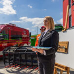 Woman in a black suit with a clipboard speaks beside a red fire department van and emergency equipment during a demonstration.