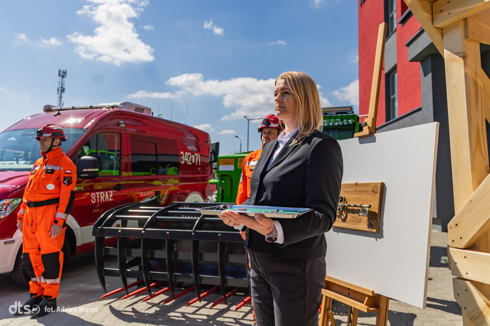Woman in a black suit with a clipboard speaks beside a red fire department van and emergency equipment during a demonstration.