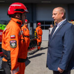 Polish firefighter in orange gear greeting a suited official outdoors beside a red fire truck.