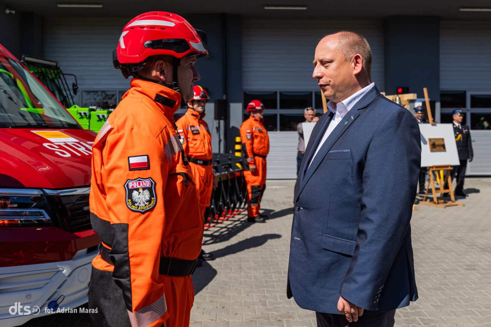 Polish firefighter in orange gear greeting a suited official outdoors beside a red fire truck.