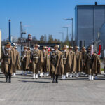A group of uniformed soldiers marching with brass instruments in an industrial outdoor yard under a blue sky.