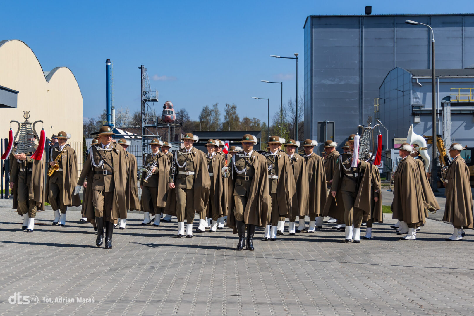 A group of uniformed soldiers marching with brass instruments in an industrial outdoor yard under a blue sky.