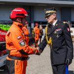 Polish emergency responder in orange gear shakes hands with a high-ranking officer in dress uniform during a ceremony, with an ambulance behind them.