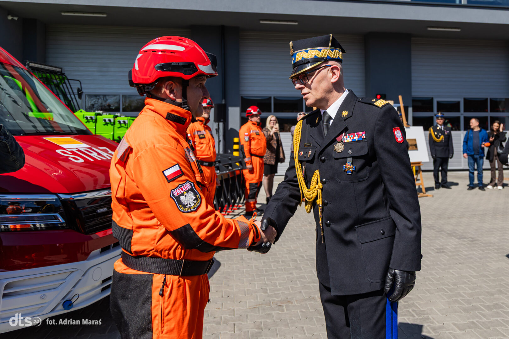 Polish emergency responder in orange gear shakes hands with a high-ranking officer in dress uniform during a ceremony, with an ambulance behind them.