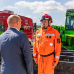 Emergency responder in bright orange suit and helmet talks to a man in a suit beside parked red fire van and green rescue vehicle behind them.