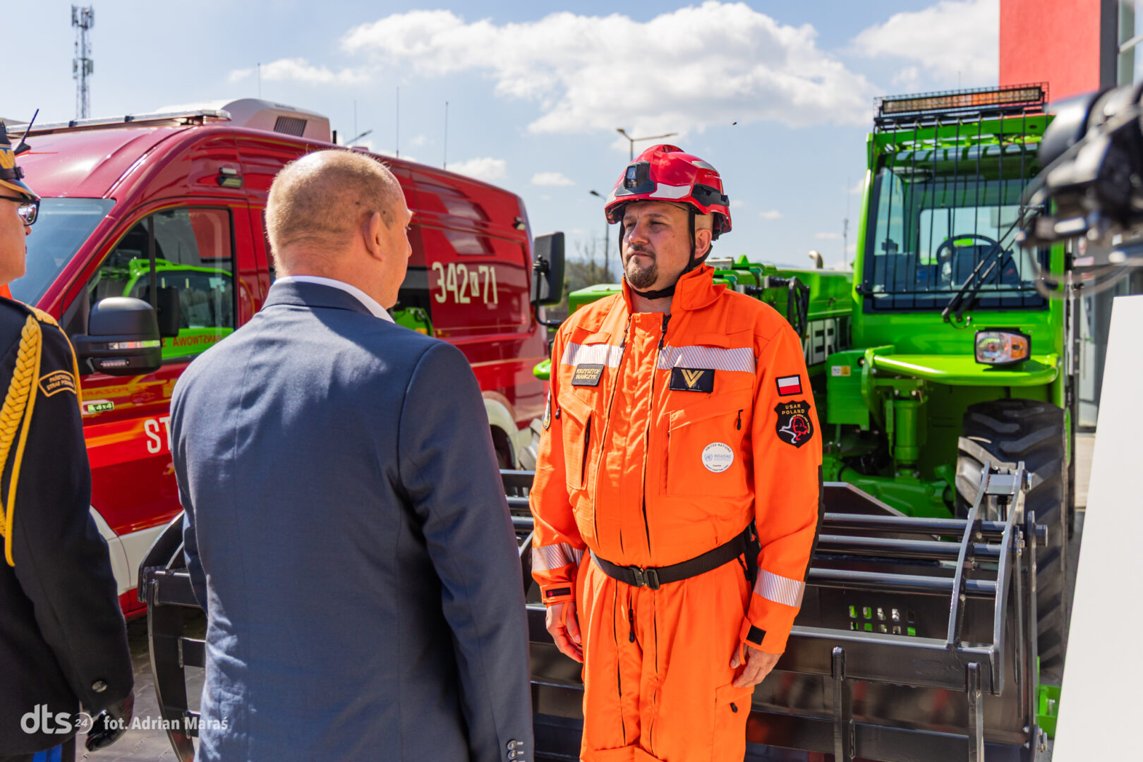 Emergency responder in bright orange suit and helmet talks to a man in a suit beside parked red fire van and green rescue vehicle behind them.